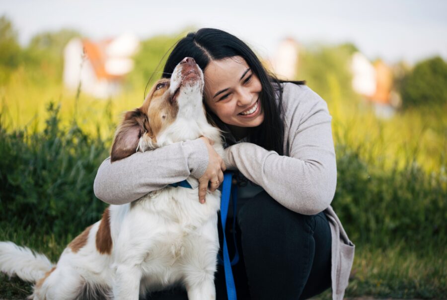 A person with long dark hair is kneeling outdoors in a grassy field, affectionately hugging a white and brown dog with a blue leash. The dog appears to be licking or nuzzling the person's face, and both seem joyful. The background is a soft blur of green grass and distant buildings under a light sky.